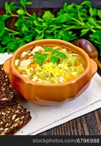 Minestrone soup with meat, celery, tomatoes, zucchini and cabbage, green peas, carrots and pasta in an earthenware bowl on a napkin, cheese and bread on a wooden boards background