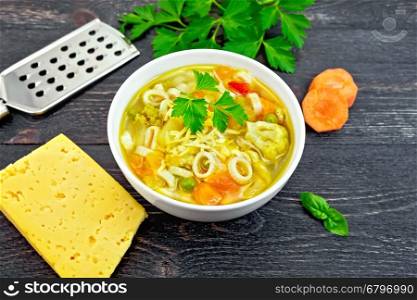 Minestrone soup with meat, celery, tomatoes, zucchini and cabbage, green peas, carrots and pasta in a white bowl, cheese, grater on background black wooden plank