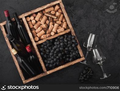 Mini bottles of red wine and empty glasses with dark grapes with corks and opener inside vintage wooden box on black stone background.