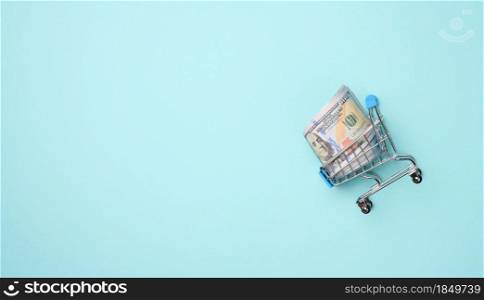 miniature shopping cart with a stack of paper American dollars on a light blue background. Savings, sale, copy space