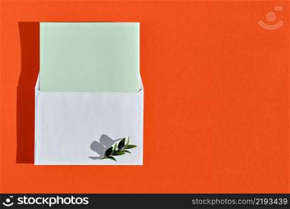 Minimal composition with white envelope, green blank card and green leaves on burnt orange background, top view with copy space. Mockup with envelope and blank card, flat lay