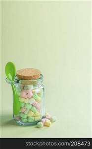 Minimal image with a glass bottle full of various colored tiny marshmallows and a green spoon attached to it, on a greenish paper background.