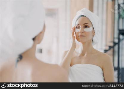 Mirror reflection of girl which applies anti wrinkle eye patches and doing skincare. Attractive caucasian woman wrapped in towel after bathing and hair washing. Relaxation at spa resort or at home.. Mirror reflection of girl which applies eye patches and doing skincare. Relaxation at home.