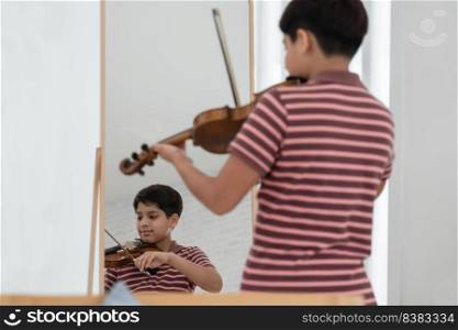Mirror reflection of Indian handsome kid boy is practicing playing wooden violin with happiness and graceful posture, standing in the mirror. Copy space. Education and talent in child concept