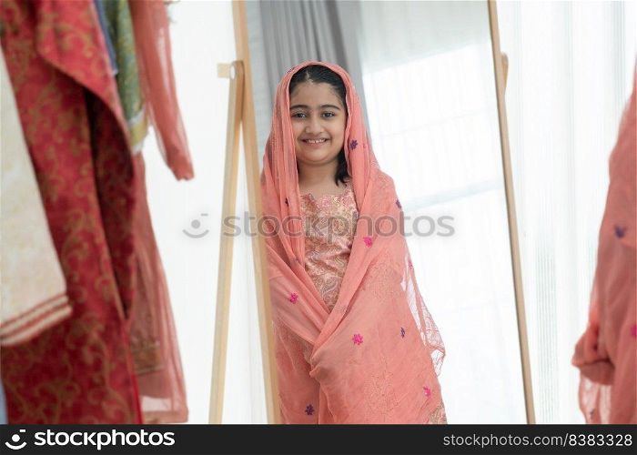 Mirror reflection of Indian little cute kid girl in traditional clothing smiling at mirror with beautiful clothes she choosed while dressing at home, looking at camera.