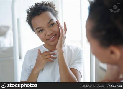 Mirror reflection of young attractive hispanic woman. Happy gorgeous african american girl applying cream to her face. Skin cleansing and treatment. Beauty routine and hygiene of tender teenage girl.. Happy african american girl is applying cream to her face. Skin cleansing and treatment.