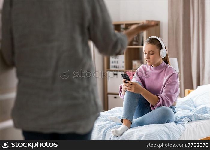 misbehavior, conflict and family concept - angry mother entering room and sad daughter in headphones with smartphone sitting on bed at home. angry mother and sad daughter with phone at home