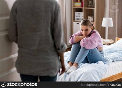 misbehavior, conflict and family concept - mother entering room and sad daughter sitting on bed at home. mother and sad daughter sitting on bed at home