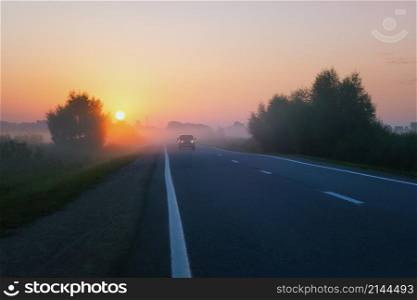 Misty golden sunrise above an suburban asphalt road in the countryside. Car with headlights in the distance drives out of the fog in the early morning at dawn.