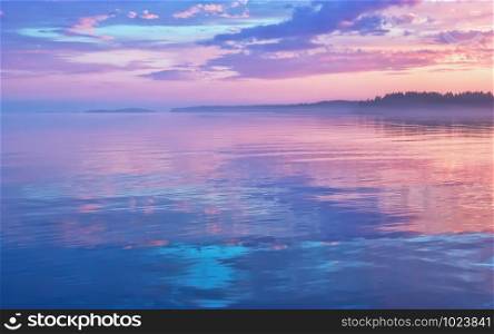 Misty seascape - calm water surface of the lake reflects lilac sky with pink and blue clouds after sunset. White nights season in the Republic of Karelia, Russia. Blur filter, space for copy.