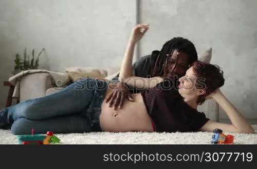 Mixed couple in love expecting baby. Happy caucasian pregnant wife and affectionate african american husband with dreadlocks lying on carpet in the living room and smiling. Loving husband caressing belly of pregnat woman and gently kissing his wife.