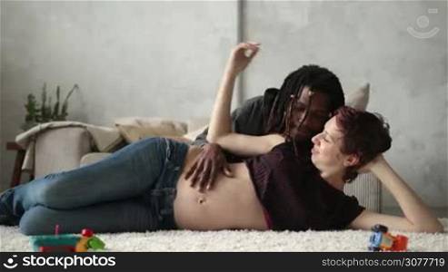 Mixed couple in love expecting baby. Happy caucasian pregnant wife and affectionate african american husband with dreadlocks lying on carpet in the living room and smiling. Loving husband caressing belly of pregnat woman and gently kissing his wife.