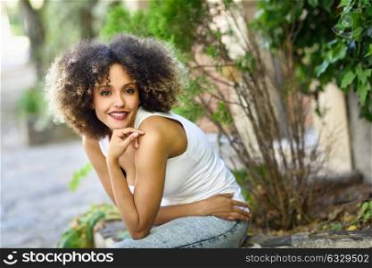 Mixed girl with afro hairstyle sitting in urban park. Young black woman wearing casual clothes.