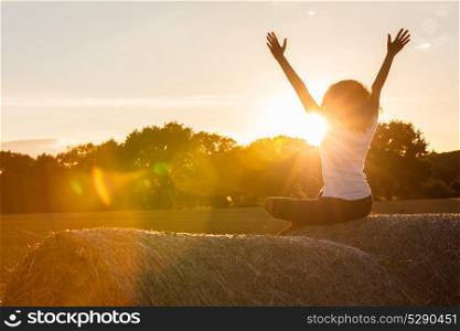 Mixed race African American girl teenager female young woman sitting on hay bale arms raised celebrating in sunset or sunrise golden evening or morning sunshine