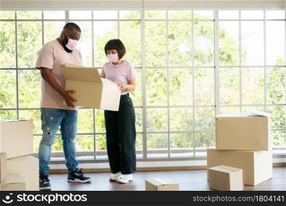 Mixed Race couple American African and Asian a surgical mask is carrying cardboard boxes in a new house on moving day. Concept of relocation, rental, and homeowner moving at home.