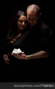 Mixed Race Couple Holding New White Baby Shoes Against a Black Background Under Dramatic Lighting.