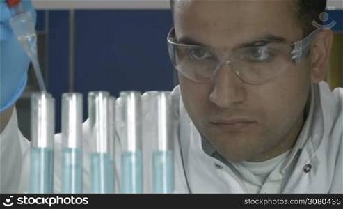 Mixed race male scientist in protective goggles holding pipette and filling test tubes with blue liquid, making research in chemical laboratory. Researcher observing the blue indicator color shift after the solution distillation in chemical lab.