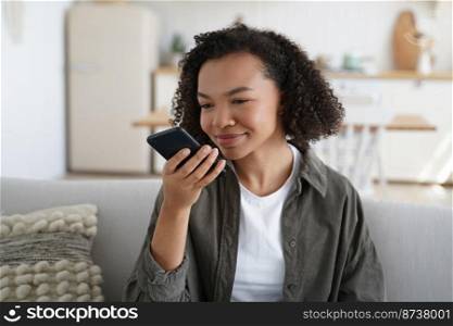 Mixed race teen girl hold phone speak voice message, chatting online, sitting on sofa. Smiling young lady uses intelligent personal virtual assistant, speakerphone or instant translation app at home.. Mixed race teen girl holding phone, speak voice message, use intelligent personal virtual assistant