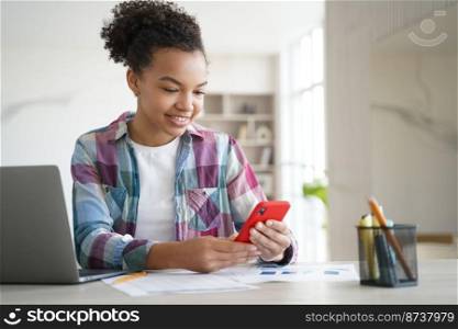 Mixed race teen girl student using smartphone, chatting in social network while learning at laptop. Schoolgirl playing online game on phone, distracted from studying doing homework. Overuse of gadgets. Mixed race teen girl student using smartphone, chatting in social network, while learning at laptop