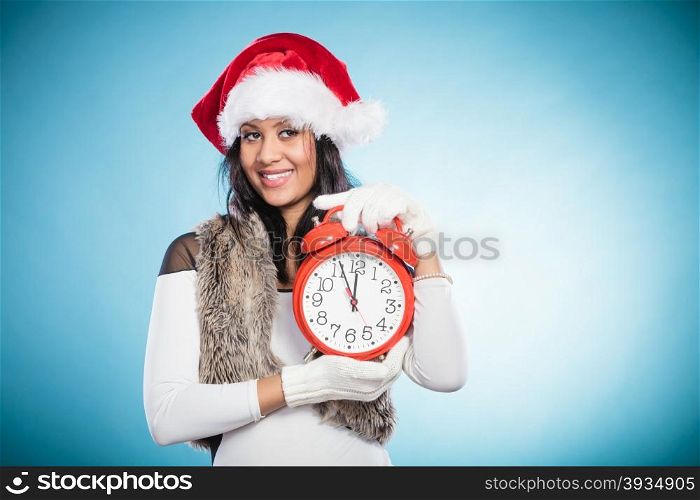 Mixed race woman in santa hat with alarm clock.. Happy cute woman in santa claus hat holding alarm clock. Mixed race african girl on blue. Christmas time season concept.
