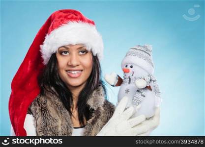 Mixed race woman in santa hat with little snowman.. Smiling cute woman in santa claus hat holding little snowman. Attractive mixed race african girl in studio on blue. Christmas season.