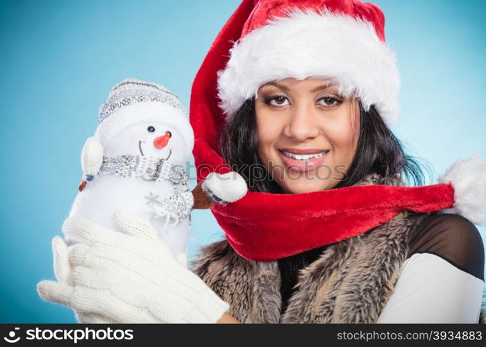 Mixed race woman in santa hat with little snowman.. Smiling cute woman in santa claus hat holding little snowman. Attractive mixed race african girl in studio on blue. Christmas season.