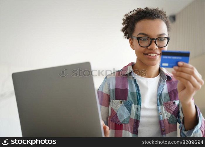 Mixed race young girl holding credit card, using online banking services at laptop, making payment. Smiling teen lady wearing glasses paying using e-bank at home. E-banking, e-commerce concept.. Mixed race teen girl holds credit card, using online banking services at laptop at home. E commerce