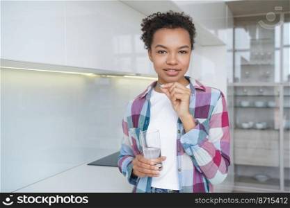 Mixed race young girl takes pills, vitamins, dietary supplement for wellness, female skin health and beauty, holding glass of water, standing at home. Vitamin deficiency prevention, healthy lifestyle.. Mixed race young girl takes pills, vitamins, dietary supplement for wellness holding glass of water