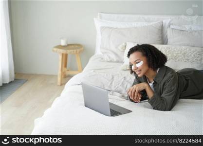 Mixed race young girl with under eye patches on face watching series on laptop lying on bed at home. Smiling pretty lady with afro hair shopping online while skincare daily routine in bedroom.. Mixed race girl use under eye patches watching series on laptop lying on bed. Skincare daily routine