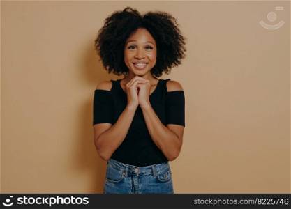 Mixed race young woman holding hands in prayer gesture, smiling with eyes opened isolated over dark beige background with copy space. African lady posing in studio, hope and positive emotions concept. Mixed race young woman holding hands in prayer gesture, smiling with eyes opened