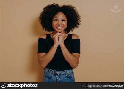 Mixed race young woman holding hands in prayer gesture, smiling with eyes opened isolated over dark beige background with copy space. African lady posing in studio, hope and positive emotions concept. Mixed race young woman holding hands in prayer gesture, smiling with eyes opened