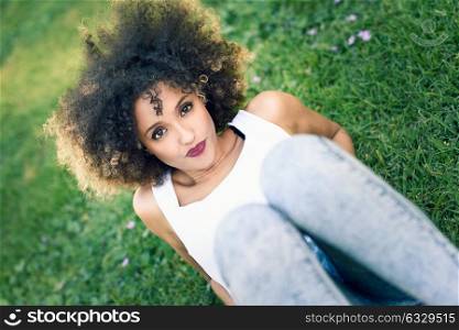 Mixed woman with afro hairstyle laying in urban park. Young black girl wearing casual clothes sitting on the grass looking at camera.