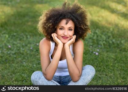 Mixed woman with afro hairstyle smiling in urban park. Young black girl wearing casual clothes sitting on the grass looking at camera.