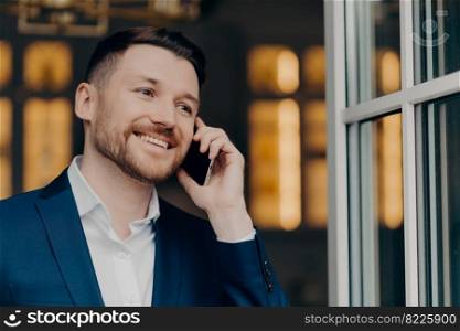 Mobile call. Handsome male executive manager with bristle has telephone conversation enjoys modern communication smiles happily looks thoughtfully into distance wers white shirt and elegant suit
