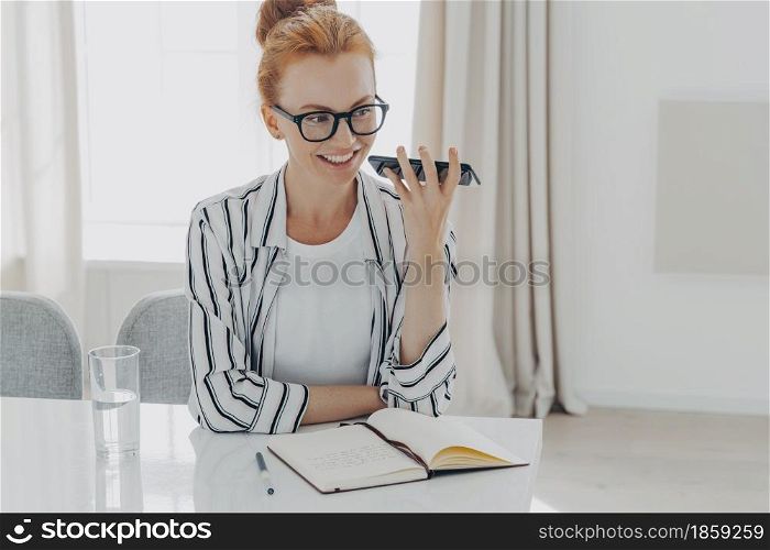 Mobile communication and voice assistant concept. Positive redhead woman uses cellphone audio recognition to send message holds gadget near mouth makes notes in diary sits at desktop drinks water. Positive redhead woman uses cellphone audio recognition to send message