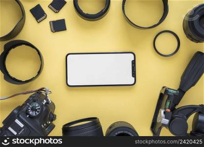 mobile phone with blank screen surrounded by modern camera accessories yellow background