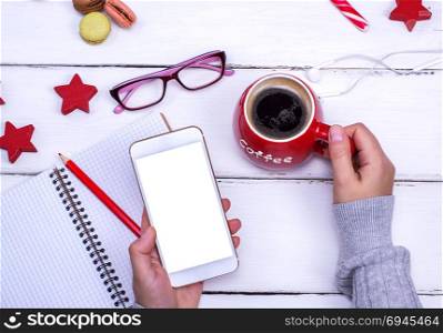 mobile phone with blank white screen in left female hand over white wooden table, in right hand red cup with black coffee, top view