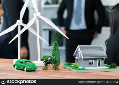 Mockup electric car with eco-friendly energy infrastructure on table with blurred background of productive business team meeting to contribute natural preservation and sustainable future. Quaint. EV car model with mockup wind turbine on blurred background meeting. Quaint