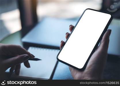 Mockup image of a woman&rsquo;s hand holding black mobile phone with blank white desktop screen with laptop and notebook on table