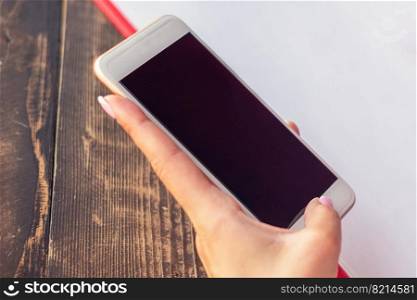 Mockup image of a woman"s hands holding white mobile phone with blank black screen on background of white sheet of paper at a cafe.. Hand of woman holding smart phone.
