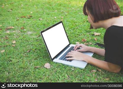 Mockup image of a woman using laptop with blank white screen lying on grass in nature outdoor park. Happy hipster young asian women working on laptop in park. Lifestyle and technology concepts.