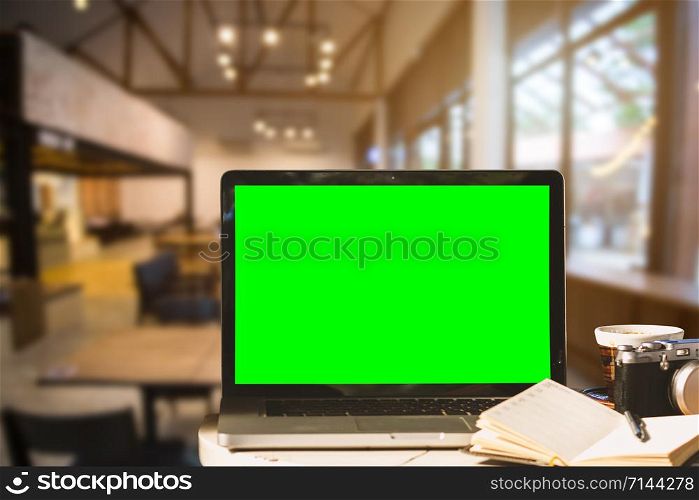 Mockup image of laptop with blank green screen with camera,notebook,coffee cup on wooden table of In the coffee shop background.