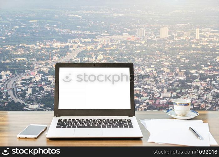 Mockup image of laptop with blank white screen,smartphone,coffee cup on wooden table view outdoors of office building dense cityscape high angle view background.