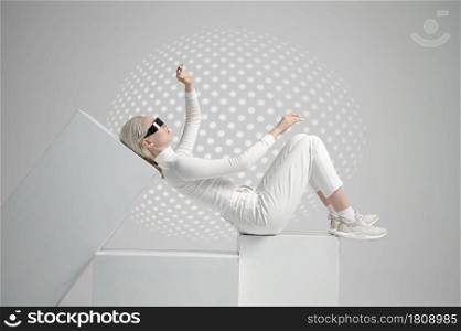 Model in white clothes and futuristic glasses lying on cubes, light grey background. Female person in virtual reality style, future technology, futurism concept. Model in futuristic glasses lying on cubes