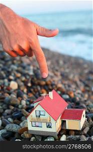 model of house with garage on stony beach in evening, Man&acute;s hand with forefinger over house
