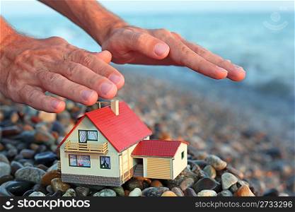 model of house with garage on stony beach in evening, Man&acute;s hands over house