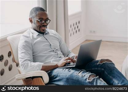 Modern african american businessman in glasses working, typing on laptop, sitting in office chair. Smiling black young guy freelancer works on business project, answering email. Remote job concept.. Modern african american businessman in glasses working, typing on laptop, sitting in office chair