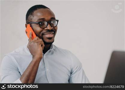 Modern african american businessman manager in glasses answers phone business call, working at laptop. Pleased black male enterpriser consulting client, talking by smartphone, using mobile connection.. Modern african american businessman manager in glasses answers phone business call working at laptop