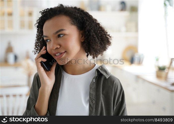 Modern african american girl talking on smartphone at home, stylish young woman with afro hairs answer mobile phone call, having conversation with friends or relatives indoors.. Modern african american young girl talking on smartphone, answering mobile phone call at home