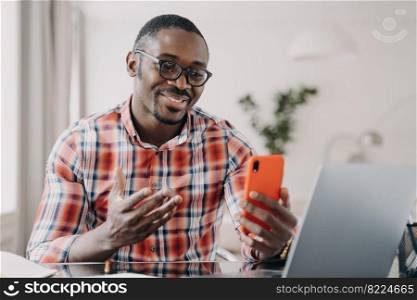 Modern african american man in glasses talking online by video call, holding smartphone, using mobile apps. Friendly smiling young black guy looking at phone screen, sitting at desk with laptop.. African american man in glasses talking online by video call, holding smartphone, using mobile apps